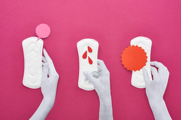 Vue du dessus des mains blanches coupées en papier et des serviettes hygiéniques blanches avec cartes et gouttes de sang sur fond violet — Photo de stock