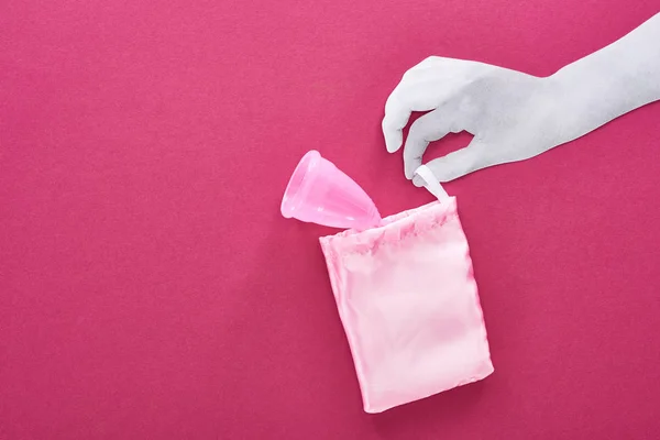 Vista superior de papel cortado mão branca com copo menstrual de plástico no saco no fundo roxo — Fotografia de Stock