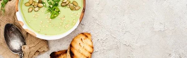 Panoramic shot of tasty green creamy soup with croutons and sprouts on textured grey background with rustic cloth — Stock Photo
