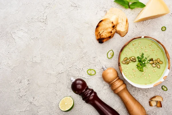 Top view of tasty green creamy soup with croutons, cheese and salt and pepper mills on textured grey background — Stock Photo