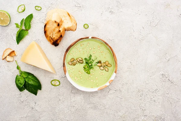 Вид сверху на вкусный зеленый сливочный суп в миске с гренками на сером фоне с текстурой — стоковое фото