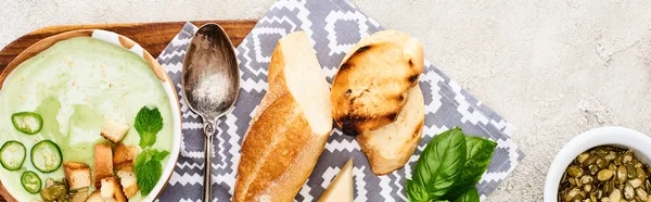 Panoramische Aufnahme grüner Gemüsecremesuppe auf hölzernem Schneidebrett mit Serviette, Löffel und Croutons in der Nähe von Kürbiskernen — Stockfoto