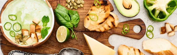 Panoramic shot of green vegetable creamy soup on wooden chopping board with ingredients on textured surface — Stock Photo