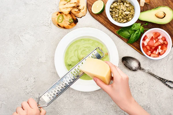 Обрезанный вид человека тертый сыр в зеленом сливочном супе возле деревянной доски резки со свежими ингредиентами — стоковое фото