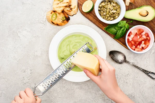 Abgeschnittene Ansicht eines Mannes, der Käse in grüner cremiger Suppe in der Nähe eines hölzernen Schneidebretts mit frischen Zutaten reibt — Stockfoto