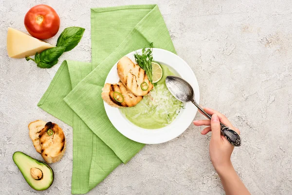 Vista cortada da mulher segurando colher perto da placa com deliciosa sopa de legumes verde cremoso com croutons — Fotografia de Stock
