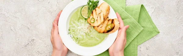 Vista ritagliata della donna che tiene piatto con deliziosa zuppa di verdure verde crema con crostini vicino tovagliolo verde, colpo panoramico — Foto stock
