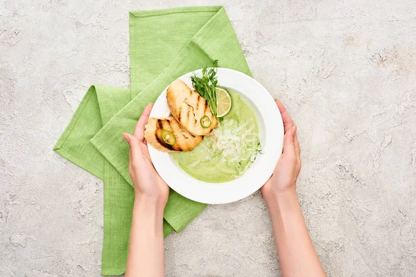 Vista ritagliata della donna che tiene il piatto con deliziosa zuppa di verdure verde crema con crostini vicino al tovagliolo verde — Foto stock