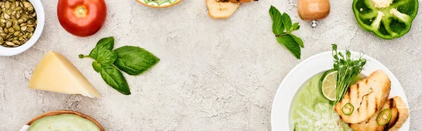 Panoramic shot of creamy green vegetable soup near fresh vegetables on textured surface — Stock Photo