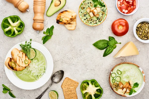 Вид на вкусный сливочно-зеленый овощной суп в мисках возле свежих овощей на текстурированной поверхности — стоковое фото
