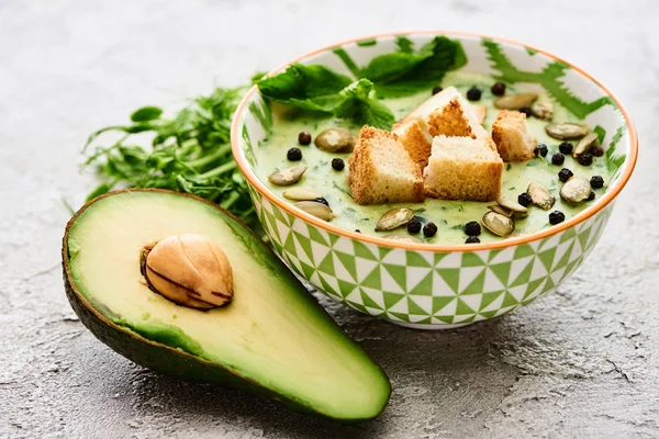 Ciotola di deliziosa zuppa verde cremosa con crostini, pepe nero e semi di zucca vicino avocado maturo — Foto stock