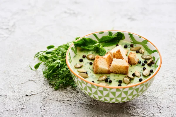 Bol de délicieuse soupe crémeuse de légumes verts avec croûtons, poivre noir et graines de citrouille près des choux verts — Photo de stock