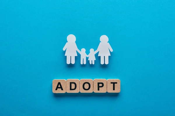 Top view of paper cut lesbian family holding hands on blue background with adopt lettering on wooden cubes — Stock Photo