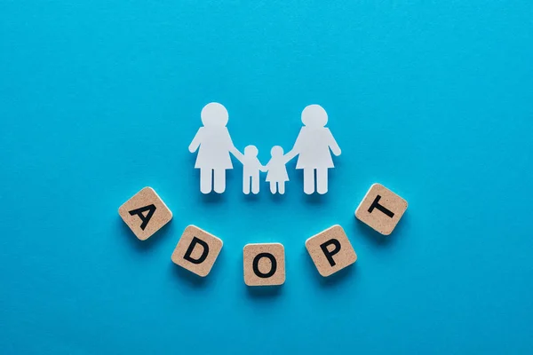 Top view of paper cut lesbian family holding hands on blue background with adopt lettering on cubes — Stock Photo