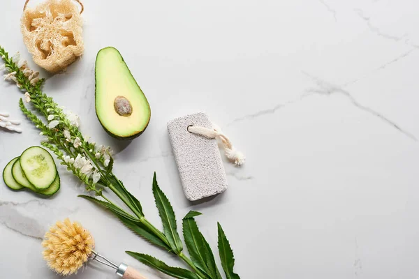 Top view of branch with flowers, fresh avocado and cucumber near body brush, loofah and pumice stone on marble surface — Stock Photo