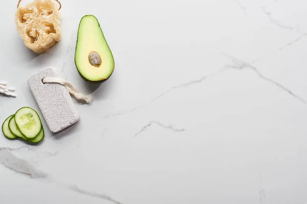 Vista superior de aguacate fresco y pepino cerca de loofah, orejeras y piedra pómez en la superficie de mármol - foto de stock