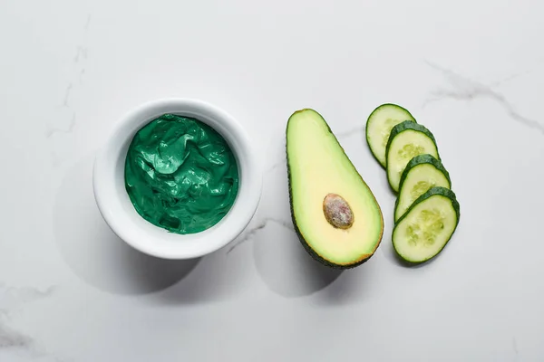 Draufsicht auf natürliche Lehmmaske in der Nähe von frischer Avocadohälfte und Gurkenscheiben auf Marmorhintergrund — Stockfoto