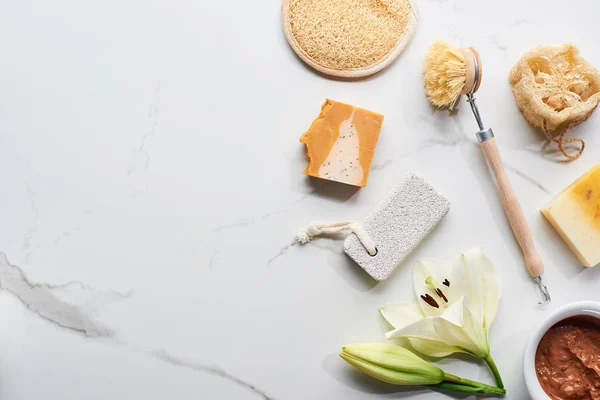 Vista superior da máscara de barro natural, peças de sabão, escova do corpo, pedra-pomes e loofah perto de flor fresca na superfície de mármore — Fotografia de Stock