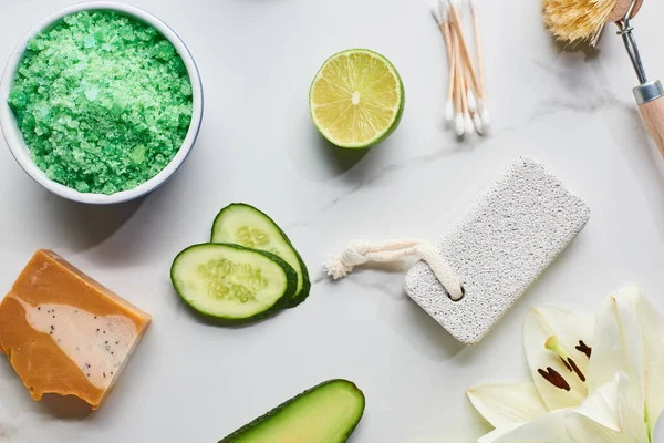 Top view of green bath salts, soap and pumice stone near fresh flower, lime and cucumber on marble surface — Stock Photo