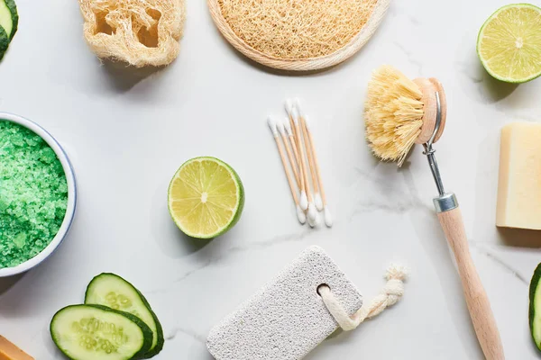 Top view of green bath salts, body brush, loofah, bath sponge and pumice stone near fresh lime and cucumber on marble surface — Stock Photo