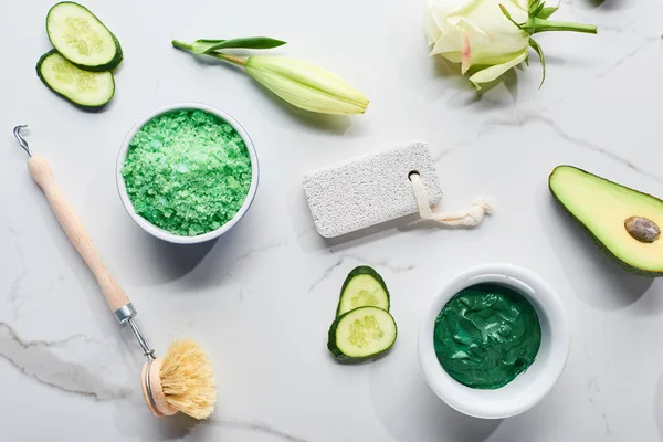 Vista dall'alto di spazzola corpo, pietra pomice, sali da bagno e maschera viso vicino a metà avocado fresco, fette di cetriolo e fiore su sfondo di marmo — Foto stock