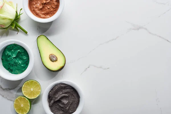 Vue de dessus des tasses avec des produits de beauté naturels près de la fleur, le citron vert et l'avocat sur la surface de marbre — Photo de stock