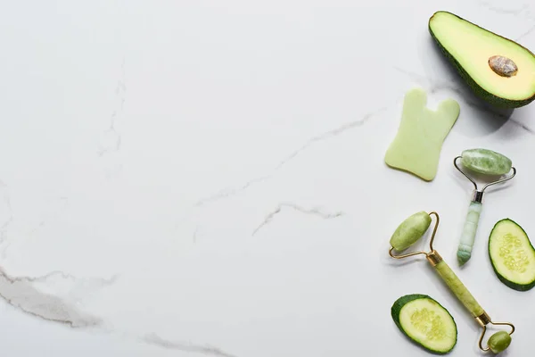 Vista superior de la espátula facial y rodillos cerca de aguacate y pepino en la superficie de mármol - foto de stock