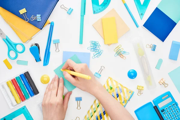 Top view of schoolgirl writing on blue card with yellow pen near stationery isolated on white — Stock Photo
