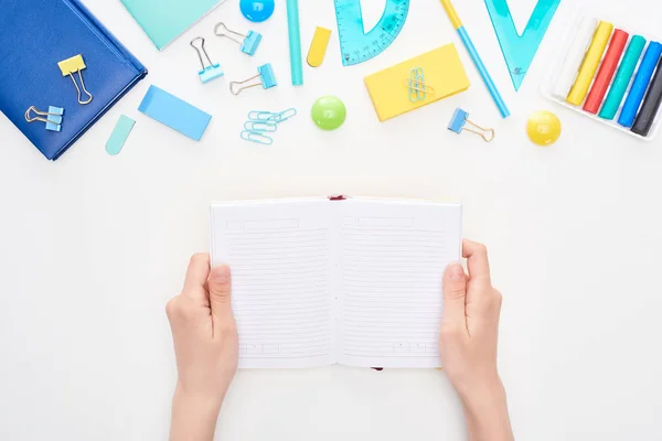 Top view of schoolgirl holding opened notebook with clear pages near stationery isolated on white — Stock Photo