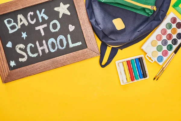 Kreidetafel in Holzrahmen mit Schulrückentafel in der Nähe blauer Schultasche und Aquarellfarben auf gelbem Hintergrund — Stockfoto