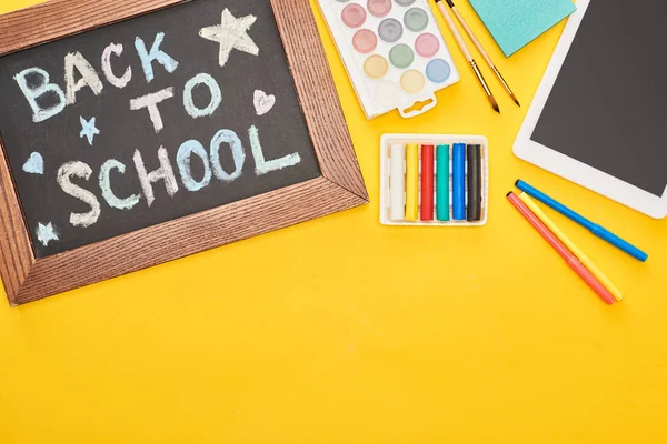 Kreidetafel in Holzrahmen mit Schulrückenbeschriftung in der Nähe von Schulmaterial und digitalem Tablet auf gelbem Hintergrund — Stockfoto