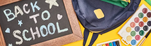 Panoramic shot of chalkboard in wooden frame with back to school inscription near blue schoolbag and watercolor paints set on yellow background — Stock Photo