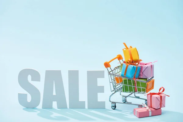 White sale lettering and festive gift boxes in shopping cart on blue background — Stock Photo