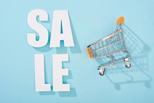 Top view of white sale lettering and decorative shopping cart on blue background — Stock Photo