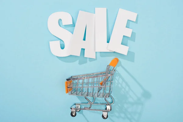 Top view of white sale lettering and empty shopping cart on blue background — Stock Photo