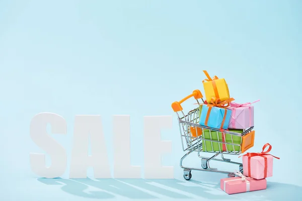 White sale lettering and festive gift boxes with bows in shopping cart on blue background — Stock Photo