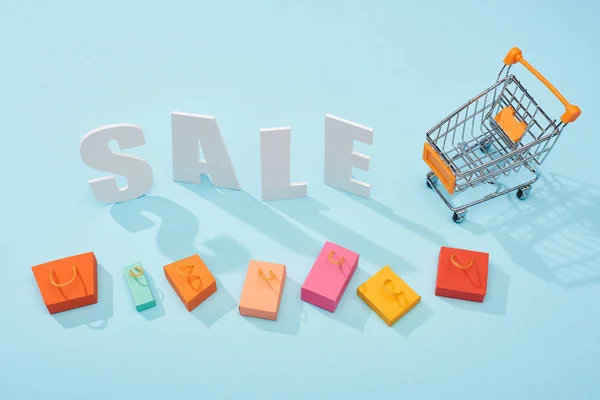 Top view of white sale lettering near shopping trolley and colorful shopping bags on blue background — Stock Photo