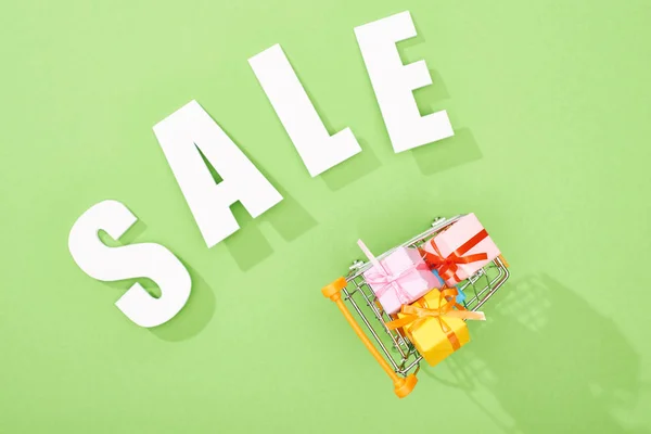 Top view of gift boxes in shopping cart near white sale lettering on green background — Stock Photo