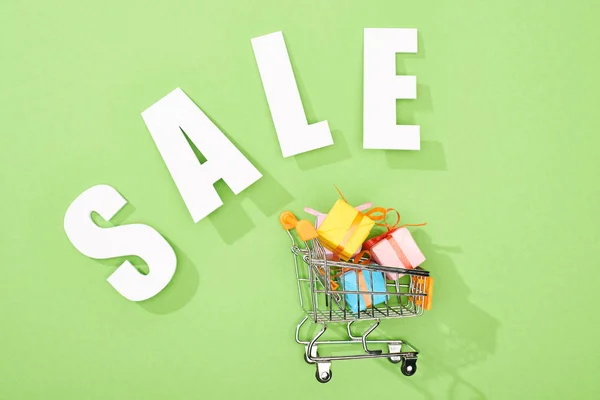 Top view of decorative gift boxes in shopping cart near white sale lettering on green background — Stock Photo