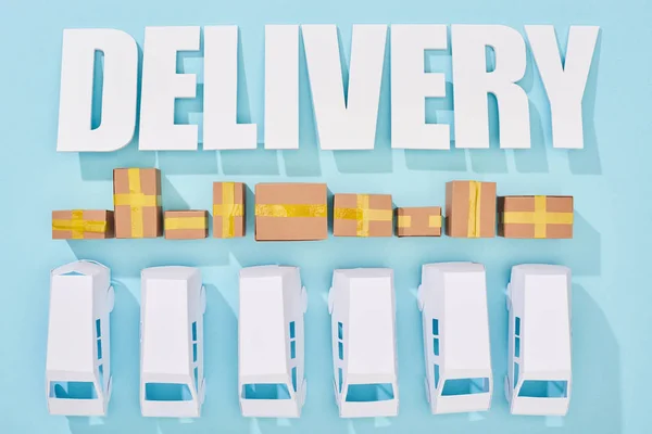 White delivery inscription with shadows over cardboard boxes and mini vans on blue background — Stock Photo