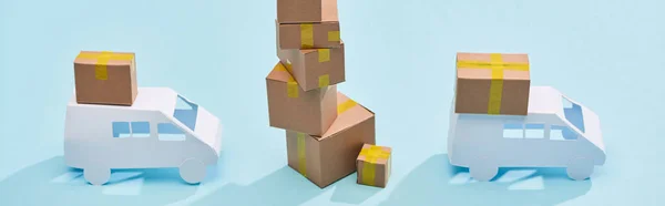 Panoramic shot of cardboard boxes one on each other near mini trucks on blue background — Stock Photo