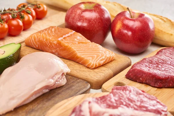 Aves de capoeira, peixe, carne, maçãs, ramo de tomate cereja, metade de abacate e baguete francês em tábuas de corte de madeira — Fotografia de Stock