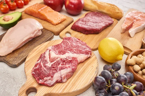 Fresh raw meat, poultry, fish on wooden cutting boards near lemon, grapes, apples, branch of cherry tomatoes, nuts and french baguette on marble surface — Stock Photo