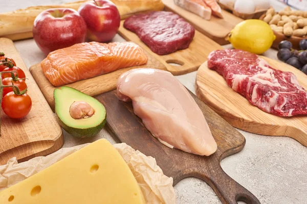 Aves frescas crudas, carne, pescado en tablas de cortar de madera cerca de manzanas, limón, queso, tomates cherry y la mitad de los aguacates - foto de stock