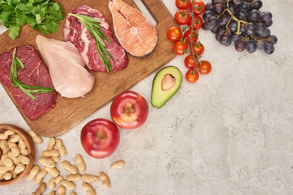 Vista superior de carne variada, aves e peixes com vegetação em tábua de corte de madeira perto de maçãs, uvas, amendoins, tomates cereja e metade do abacate na superfície de mármore cinza — Fotografia de Stock