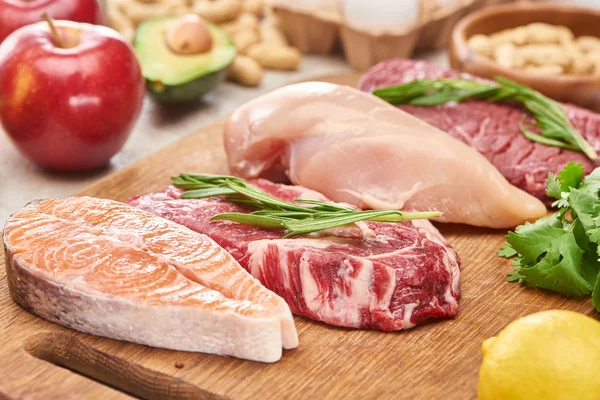 Carne variada, aves de corral y pescado con ramitas de romero sobre tabla de cortar de madera cerca de limón, manzana y aguacate - foto de stock