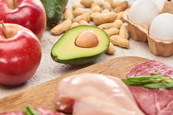 Assorted meat and poultry with rosemary twigs on wooden cutting board near apples, avocado, peanuts and eggs — Stock Photo