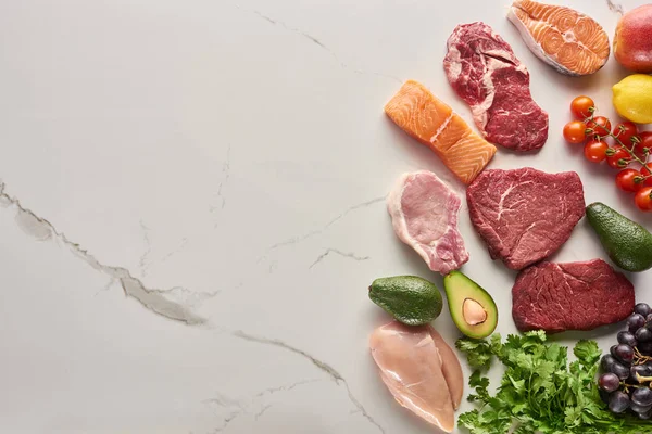 Top view of assorted meat, poultry and fish near parsley, grapes, cherry tomatoes, avocados, apple and lemon on gray marble surface — Stock Photo