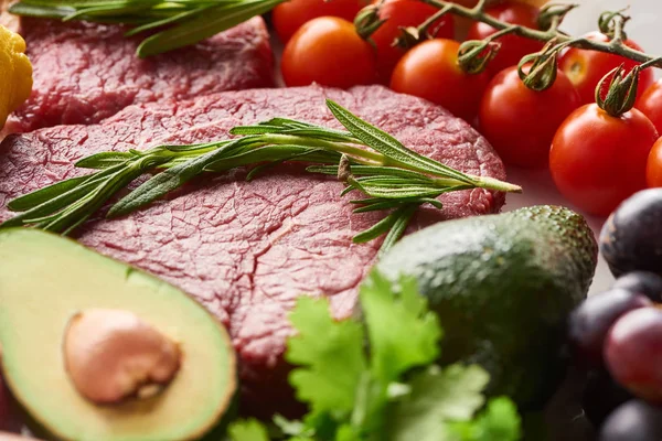 Close up view of raw meat steak with rosemary twig near avocados, branch of cherry tomatoes and parsley — Stock Photo