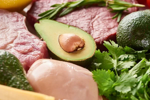 Close up view of half of avocado between raw meat and poultry with rosemary and parsley twigs — Stock Photo